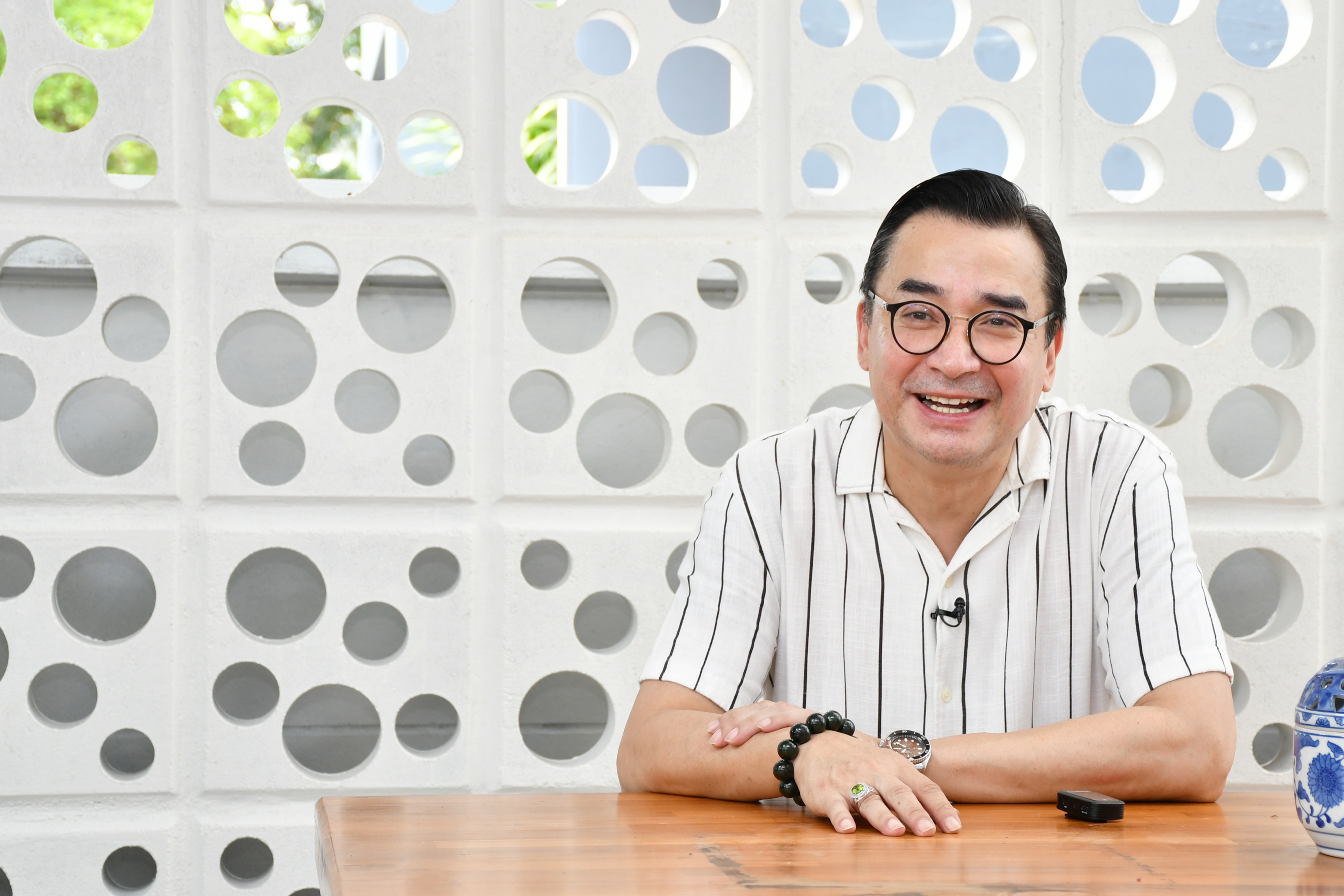 บุคคลนั่งอยู่หลังโต๊ะไม้ในอาคาร ฉากหลังเป็นผนังสีขาวลายช่องวงกลม